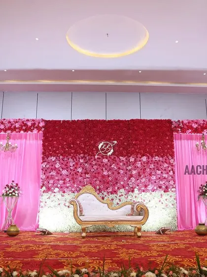 This reception stage features a beautiful ombre flower wall, transitioning from deep red to pink and finally to white. The design is framed by elegant pink drapes and chandeliers, creating a romantic and colorful setting for the couple.