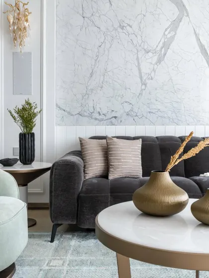A close-up of the living room's seating area, featuring a plush tufted charcoal sofa against a backdrop of intricate wall moulding and a striking marble panel. The space is styled with sculptural vases and soft textiles.