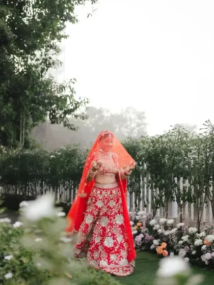 The bride stands veiled in a misty garden, creating an ethereal and mysterious portrait. The vibrant red of her lehenga pops against the soft green and white background.