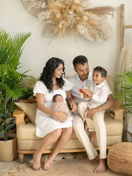 A full family portrait in a cozy, plant-filled studio setting. The older brother's excitement is balanced by the parents' calm embrace of their new baby.