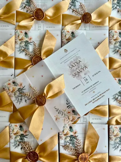 A beautiful floral wedding invitation for Satya and Sravya, featuring a vellum wrap and a luxurious gold satin ribbon. The design is soft, romantic, and perfect for a classic wedding celebration.