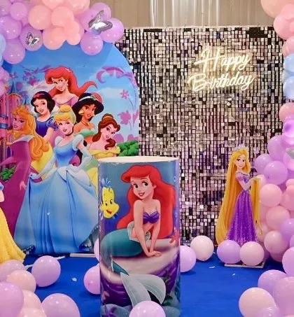 A classic Disney Princess birthday party. The setup features a backdrop with multiple princesses, a silver shimmer wall, and a custom-printed pedestal with Ariel.
