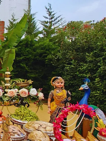 A detailed view of a traditional ceremony setup, featuring a classic South Indian doll next to a peacock figure. The space is filled with fresh fruits, flowers, and dry fruits as offerings, reflecting our attention to cultural details.