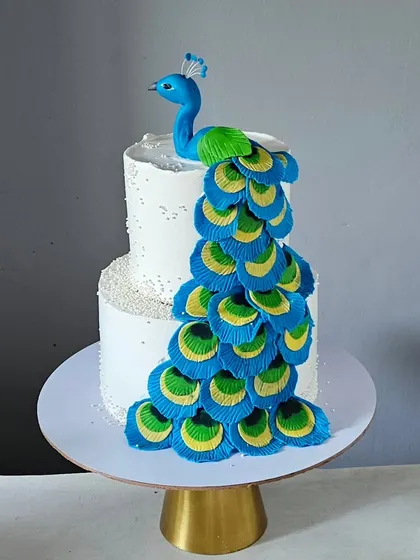 A majestic peacock-themed cake. The centerpiece is a handcrafted, edible peacock whose vibrant, cascading tail feathers are made from individual pieces of fondant, creating a true work of art.