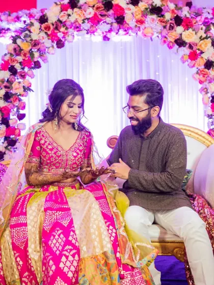 A sweet moment between the couple during their engagement ceremony. The stage is decorated with a beautiful floral arch in shades of pink and purple, creating a soft and romantic atmosphere.