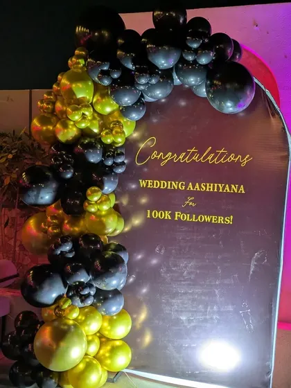 A straight-on view of the "100k Followers" celebration backdrop. The lighting from below highlights the metallic sheen of the gold balloons and the crisp text on the banner.