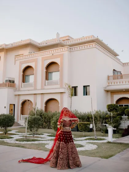 The bride in her stunning red lehenga poses in the gardens of a grand palace, her long veil trailing behind her.