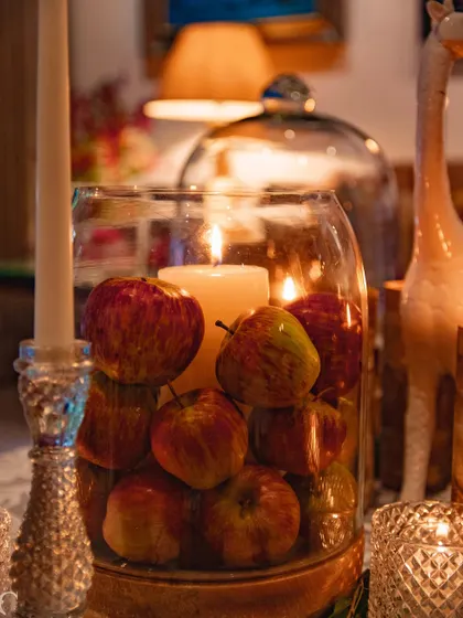 The soft, warm glow of the candle illuminates the apples from within the glass vase, creating a cozy and inviting detail on the table.