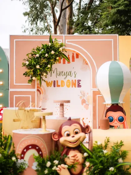 A detailed shot of the 'Wild One' backdrop, showcasing the custom signage, a whimsical hot air balloon, and a cheeky monkey prop. The soft, warm colors give this jungle theme a gentle, boho feel.