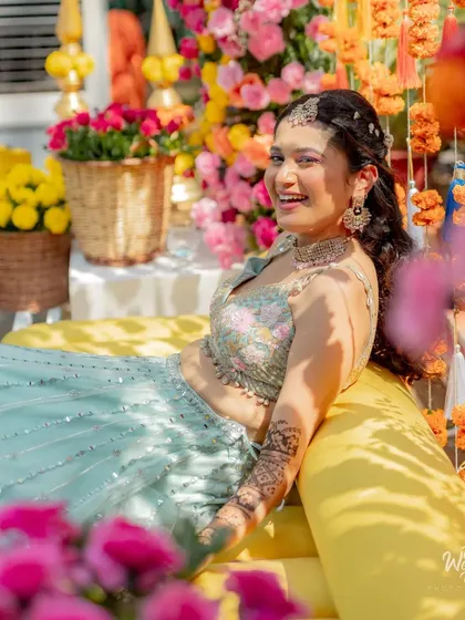 The bride enjoying her Mehendi ceremony, lounging on a bright yellow sofa surrounded by a profusion of colorful flowers.