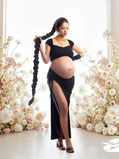 A confident and modern maternity portrait. The client's bold pose, chic black outfit, and long braided hair create a powerful, high-fashion image, beautifully framed by soft flowers.