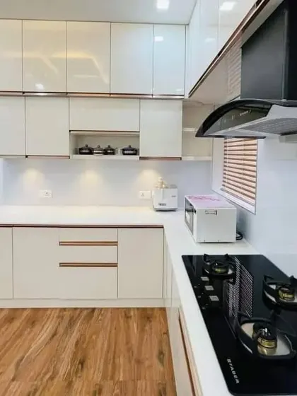 A beautiful L-shaped modular kitchen with glossy white cabinets and rose-gold profile handles. The wooden flooring adds a touch of warmth to the otherwise contemporary design.