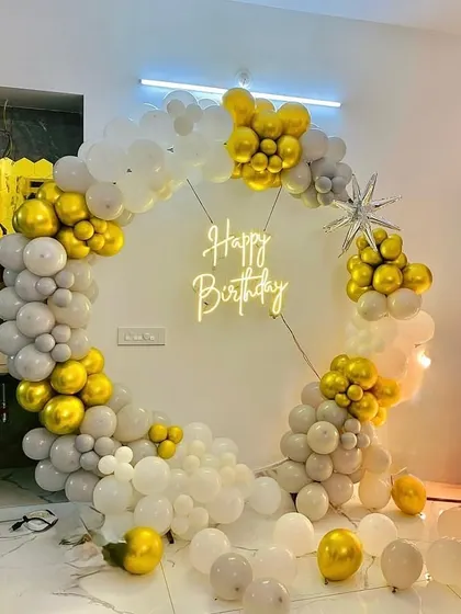 A classy birthday photo corner with a circular balloon arch in grey, white, and gold. The 'Happy Birthday' neon sign in the center makes it a perfect spot for pictures.