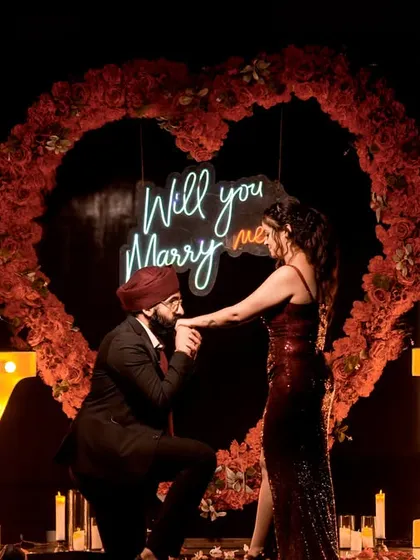 The perfect moment. He gets down on one knee in front of a heart-shaped floral arch and a neon "Will you Marry me?" sign.