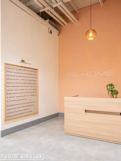 The reception area at FA Home is clean and minimal, with a soft peach accent wall and a simple wooden desk. A large framed textile piece tells the brand's story, immediately connecting customers to its artisanal roots.