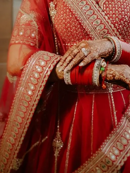 A detailed shot focusing on the bride's hands and the mid-section of her red lehenga, highlighting the intricate embroidery and her bridal choora.