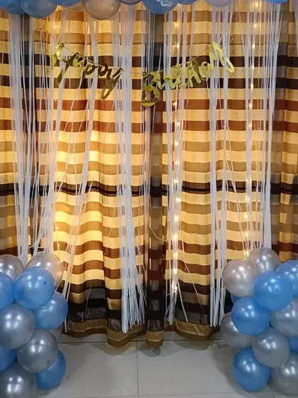 A simple yet beautiful birthday decoration with blue and silver balloon bunches framing a curtain backdrop with fairy lights. This shows how even a small space can be made festive and special with the right balloon arrangement.