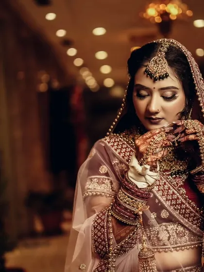 A classic bridal pose, with the bride looking down at her hands, showcasing her elaborate lehenga and jewelry.