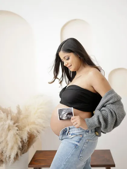 A modern and casual portrait. The mom-to-be holds her sonogram picture against her bare bump, dressed in jeans and a cozy cardigan.
