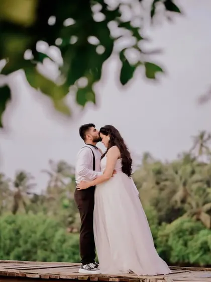 An intimate and tender pre-wedding moment. The groom-to-be kisses his fiancée's forehead, highlighting her elegant white gown and his classic suspenders look against a backdrop of palm trees.