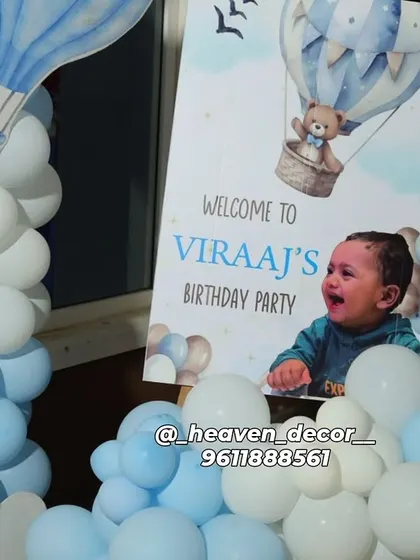 A personalized welcome sign for a hot air balloon themed first birthday. The sign includes a photo of the birthday boy and is placed next to a blue and white balloon column.