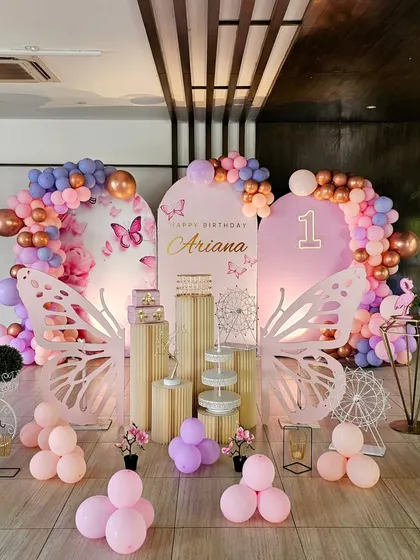 A beautiful butterfly-themed first birthday. The large, laser-cut butterfly wings create a stunning centerpiece for photos, complemented by a soft pink and purple balloon arrangement.