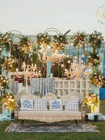An elegant outdoor Mehendi Sangeet setup. The stage, adorned with chandeliers and white florals against a blue and white patterned backdrop, created a sophisticated space for performances.