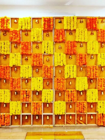 A creative and traditional backdrop featuring a grid of niches, each filled with vibrant marigold flowers and a small brass bell. This design adds a beautiful visual and auditory element to any puja or festive event.
