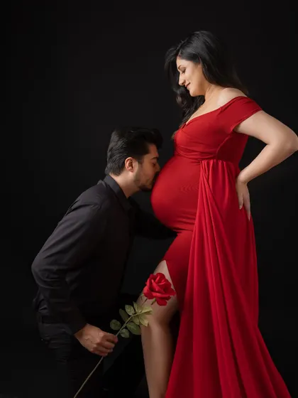 A truly romantic moment from a maternity session. The father-to-be kneels to gently kiss his partner's belly, holding a single red rose. This dramatic studio portrait with a black background highlights their love and excitement.
