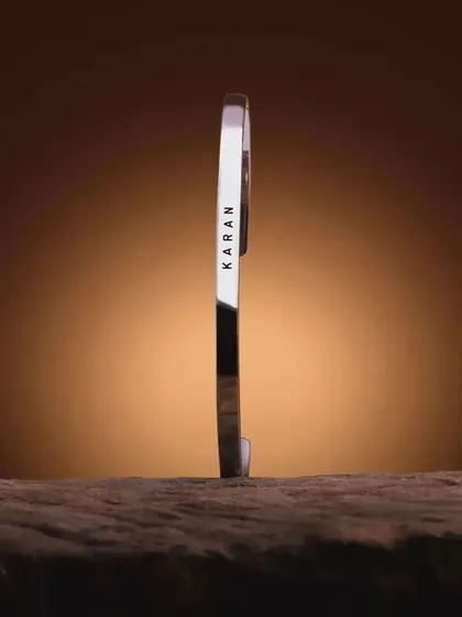 A silver cuff bracelet engraved with 'KARAN', standing vertically on a wooden surface. This artistic shot emphasizes the bracelet's solid construction and sharp engraving.