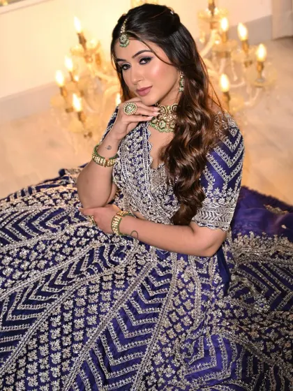 A regal seated portrait showcasing the full festive ensemble. The deep blue lehenga and dramatic makeup create a look that is fit for a modern queen.
