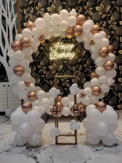 A minimalist and elegant birthday setup. A circular frame is decorated with white and rose gold balloons, with a 'Happy Birthday' neon sign in the middle.