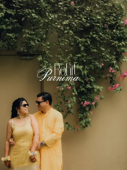 A stylish portrait of a couple in coordinated yellow attire, posing against a wall covered in beautiful bougainvillea during their Haldi event.