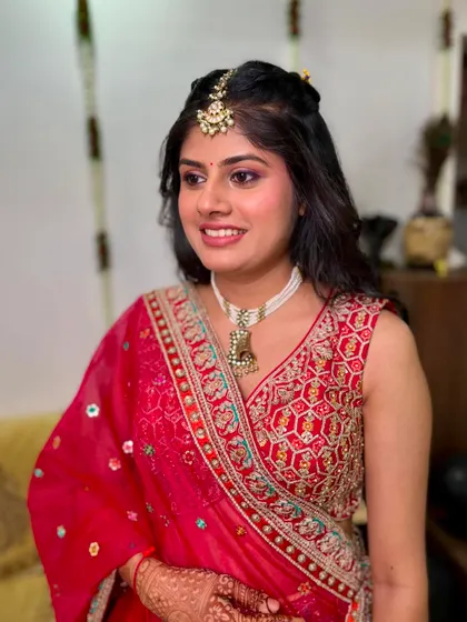 A beautiful, soft glam look for a bride in a red and pink lehenga. The focus is on rosy cheeks and shimmery eyes, creating a youthful and romantic bridal style.