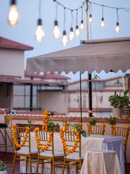 A simple and elegant rooftop setup for an intimate Haldi or Mehendi. The chairs are adorned with fresh marigold garlands, and string lights add a warm glow to the evening.