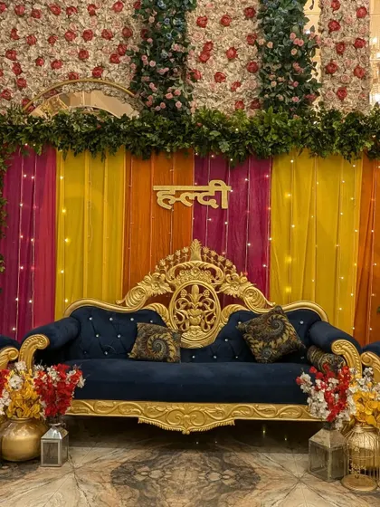 A close-up of the royal Haldi stage. The intricate gold details of the sofa and the vibrant colors of the drapes and 'Haldi' sign create a luxurious feel.
