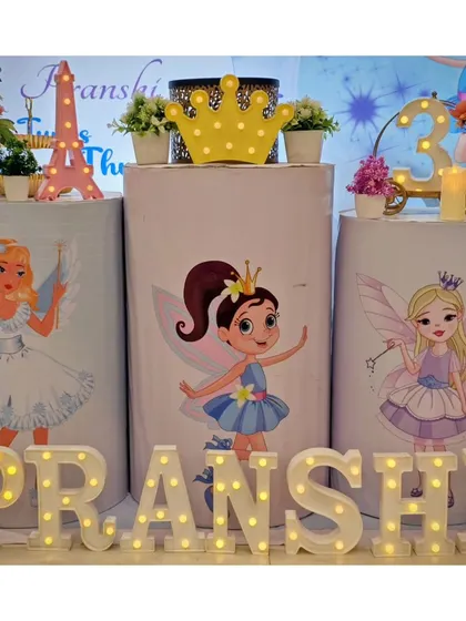 A close-up of the fairy-themed cake table, with plinths featuring different fairy character's and the name 'PRANSHI' in marquee letters.