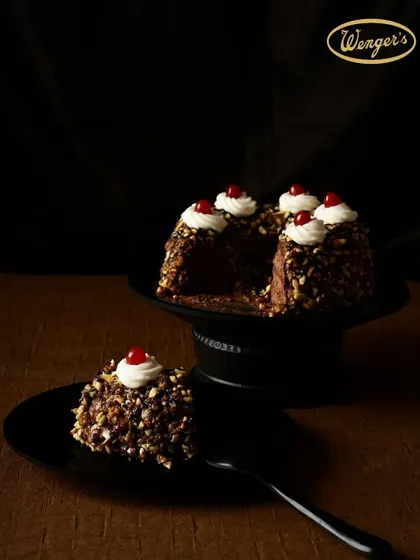 This is our Chocolate Hazelnut Cake, presented with a slice cut out to show the rich interior. It's covered in crunchy, toasted almond pieces and topped with dollops of cream and a single cherry.