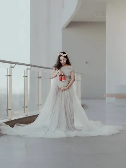 A full-length portrait of a pregnant woman in a beautiful, flowing gown, holding a pair of baby shoes. The bright, airy setting provides a perfect backdrop for this classic maternity photo.
