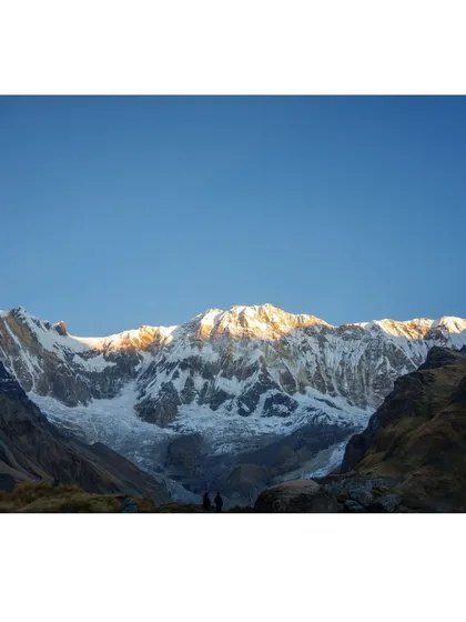 Two tiny figures of trekkers stand in silhouette against the massive, sunlit face of the Annapurna range, illustrating the incredible scale of the Himalayas.