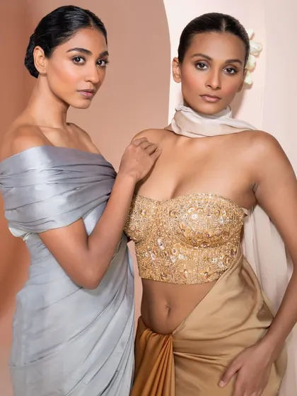 A campaign shot featuring two models in modern draped gowns. The contrast between the cool grey and warm gold creates a visually striking image.