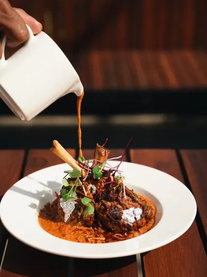 An action shot of gravy being poured over a lamb shank. The natural sunlight creates a dramatic highlight on the pour, adding a dynamic and luxurious feel to this signature dish.