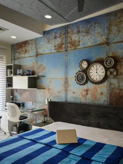 This bedroom in the Gupta Residence has an industrial-modern character. A textured feature wall resembling weathered metal panels is highlighted by a cluster of gear-inspired clocks, and a compact study corner makes the space stylish and functional.