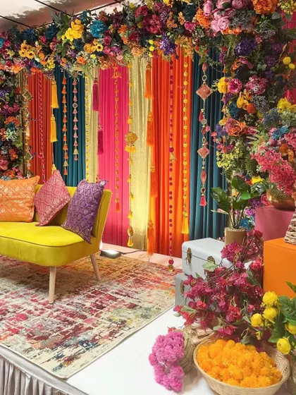A vibrant stage setup for a Mehendi ceremony. The decor features a bright yellow sofa, colorful drapes in orange, pink, and teal, and an abundance of fresh flowers.