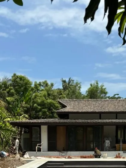 A view of a weekend home under construction in Alibaug, with the pool in the foreground. The design features a low-slung profile and a tiled roof that respects the scale of the surrounding landscape.