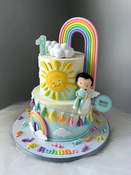 "You are my sunshine" themed first birthday cake. This cheerful two-tiered cake is decorated with a smiling sun, rainbows, and a cute, handcrafted baby figurine.