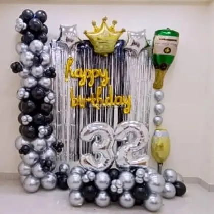 A 32nd birthday home decoration with a silver fringe backdrop. The setup includes black and silver balloon columns, a crown foil balloon, and a champagne bottle balloon.