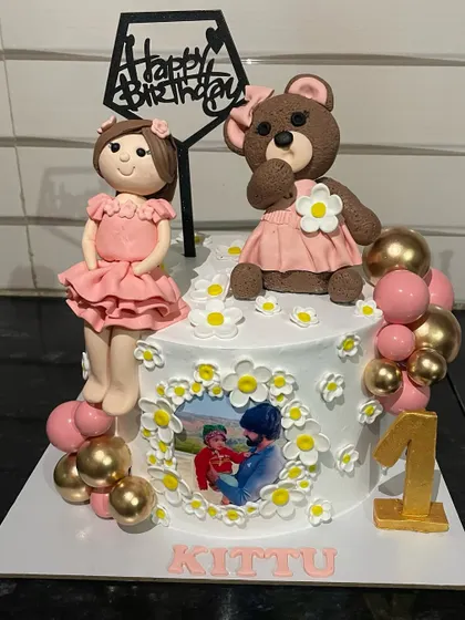 This first birthday cake is full of personal touches, featuring handcrafted fondant figures of a little girl and her teddy bear. It also includes a small, edible photo, combining a fun theme with a cherished memory.