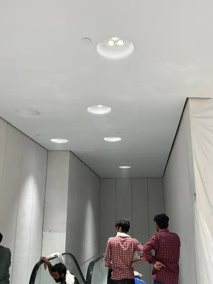 Workers completing the ceiling and lighting installation around the escalator area. The precise placement of recessed lighting is critical for achieving the clean, minimalist aesthetic required in modern retail design.
