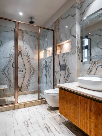 This bathroom showcases stunning book-matched marble with dramatic veining. The rose-gold frame of the shower and the textured wooden vanity add warmth and a touch of color to the luxurious space.